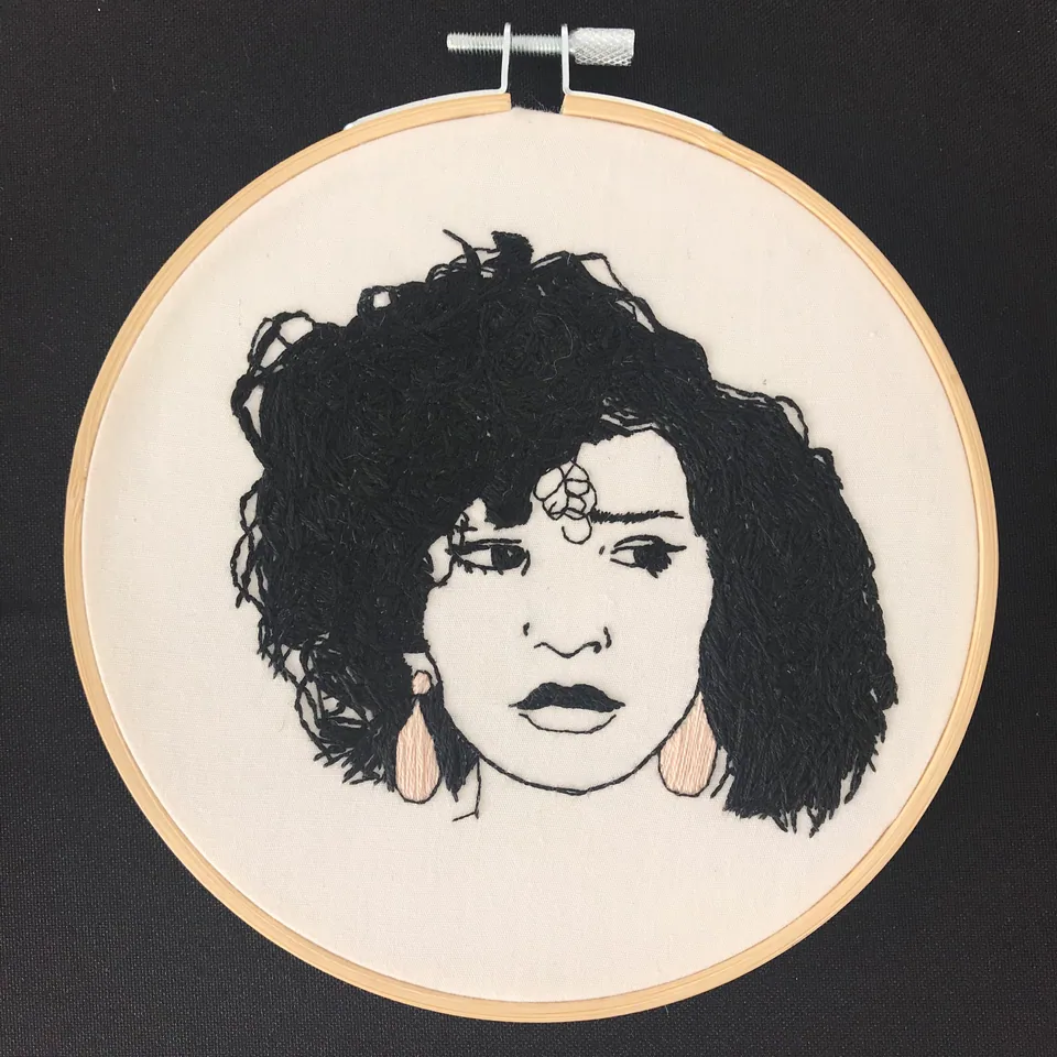 Ruida, a black-yarn portrait with pink earrings in an embroidery hoop